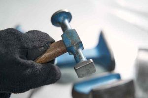 Gloved hand holding a ball-peen hammer over a metal workpiece for hand stamping