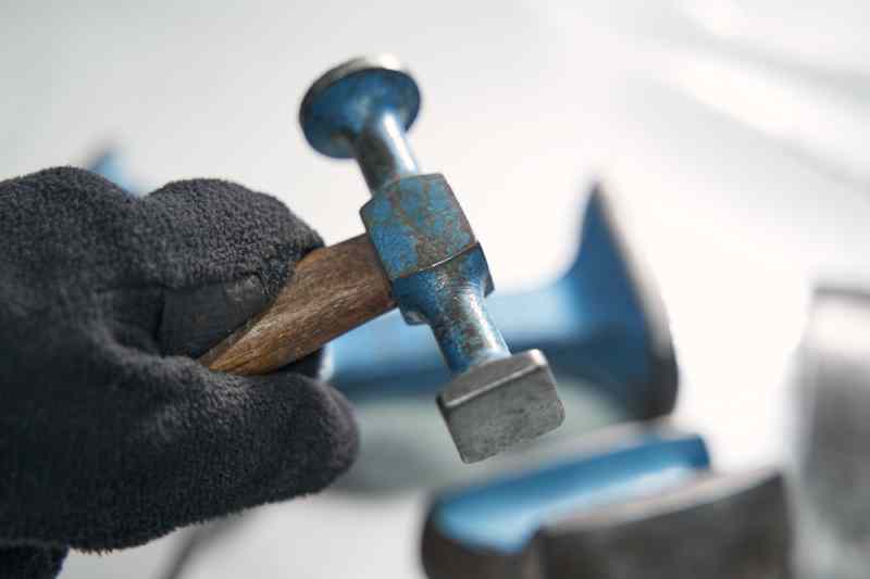 Gloved hand holding a ball-peen hammer over a metal workpiece for hand stamping